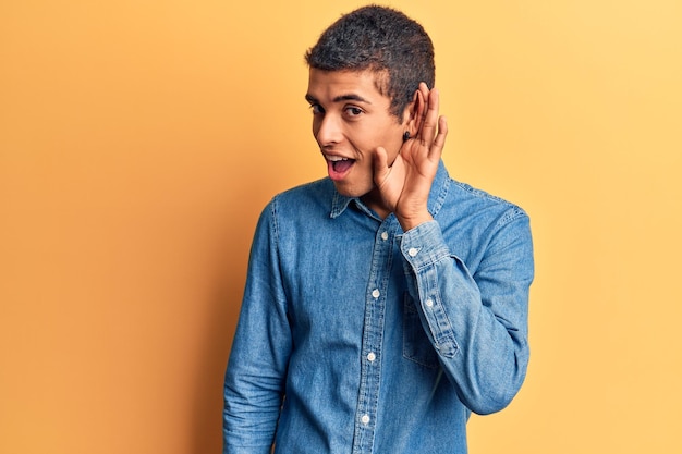 Joven afroamericano vestido con ropa informal sonriendo con la mano sobre el oído escuchando y escuchando rumores o chismes sobre el concepto de sordera