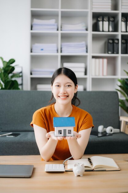 Foto joven agente inmobiliario asiático trabajando con computadora portátil y tableta en la mesa de la oficina y una pequeña casa al lado