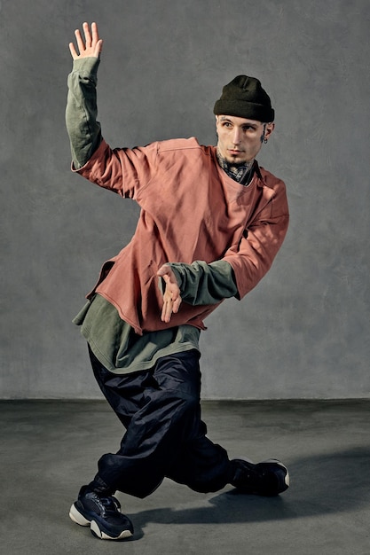 Joven agraciado con cuerpo y cara tatuados, aretes, barba. Vestido con sombrero, ropa informal y zapatillas negras. Bailando sobre fondo gris. Dancehall, hip hop. Longitud total, espacio de copia