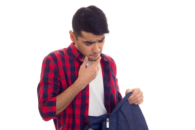 Un joven agradable con cabello negro en camiseta blanca y camisa a cuadros roja mirando su mochila
