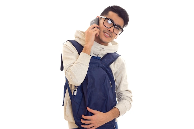 Un joven agradable con cabello negro encapuchado con gafas negras y mochila hablando por teléfono inteligente