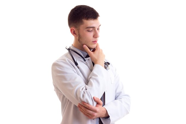 Joven agradable con camisa azul, corbata y bata de médico con estetoscopio en el cuello pensando