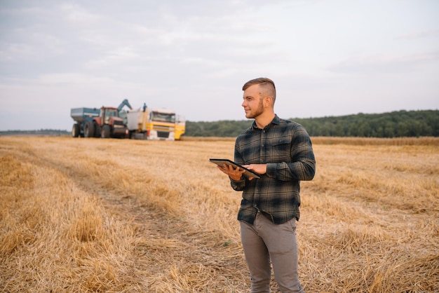 Joven agricultor atractivo con portátil de pie en el campo de trigo con cosechadora en segundo plano.