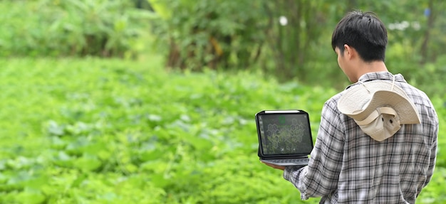 Un joven agricultor está usando una computadora portátil mientras está parado entre el huerto.