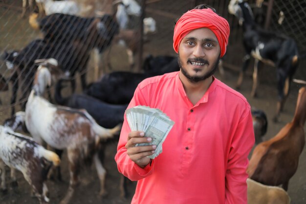 Joven agricultor indio mostrando dinero en la granja lechera de cabras