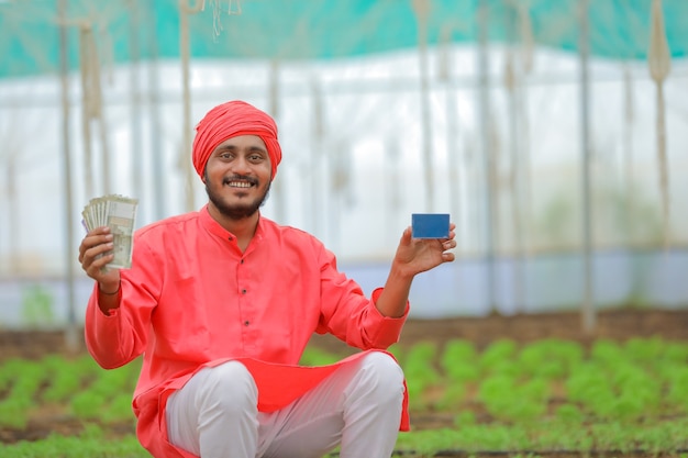 Joven agricultor indio mostrando dinero y tarjeta en blanco en invernadero