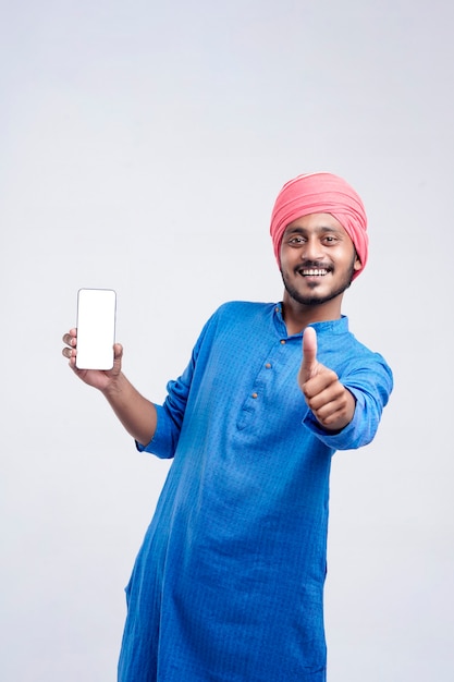 Joven agricultor indio mostrando smartphone y dando expresión sobre fondo blanco.