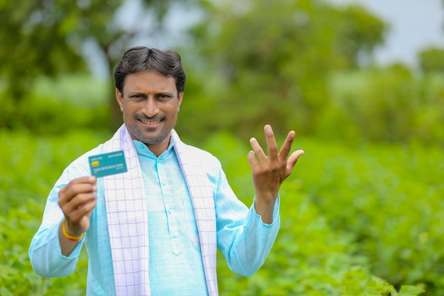 Joven agricultor indio mostrando tarjeta de débito o crédito en su campo de agricultura verde