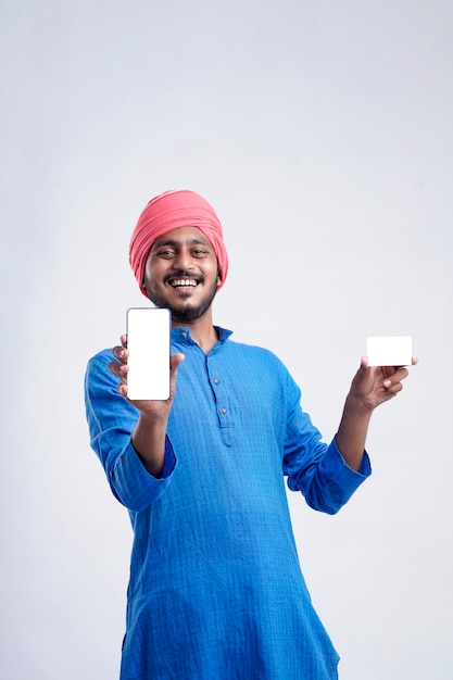 Joven agricultor indio mostrando tarjeta y smartphone sobre fondo blanco.