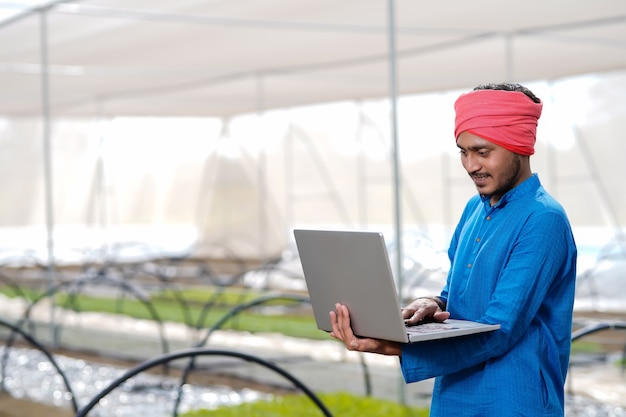 Joven agricultor indio usando laptop en invernadero o casa de polietileno