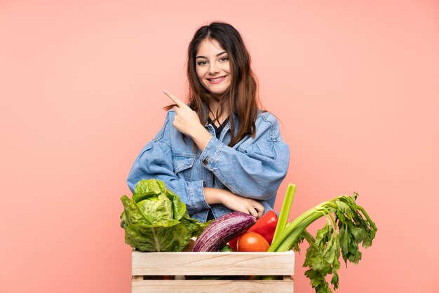 Joven agricultor mujer sosteniendo una cesta llena de verduras frescas, señalando con el dedo al lado