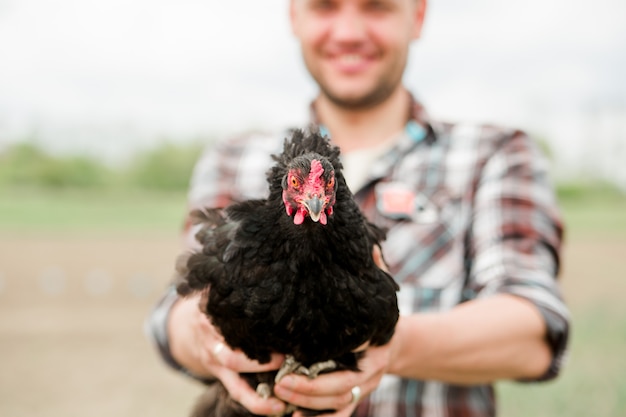 Un joven agricultor con un pollo en sus manos está parado en su jardín en el pueblo.