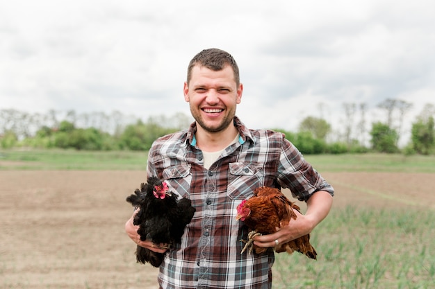 Un joven agricultor con un pollo en sus manos está parado en su jardín en el pueblo.