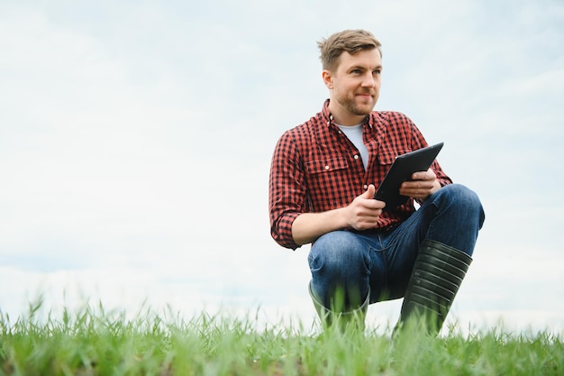 Joven agricultor sosteniendo una tableta y comprobando el progreso de la cosecha en el campo de trigo verde El trabajador sigue las perspectivas de crecimiento Concepto agrícola