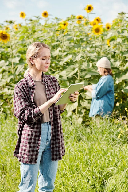 Joven agricultora seria en ropa de trabajo de pie delante de la cámara y con tableta digital contra el campo de girasol y mujer madura trabajando