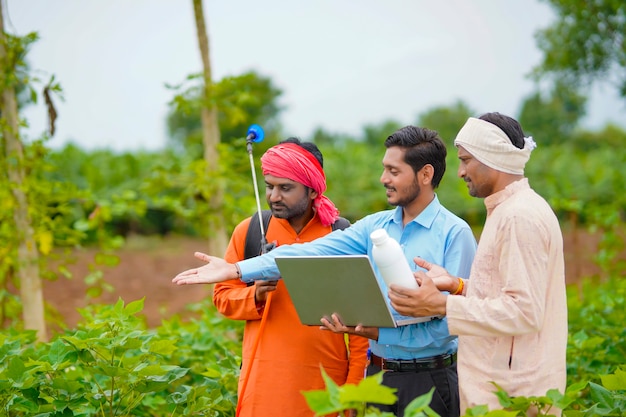 Joven agrónomo indio dando una botella de fertilizante líquido al agricultor y mostrando la información del producto en la computadora portátil en el campo de la agricultura verde.