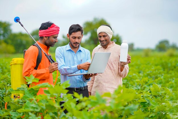 Joven agrónomo indio dando una botella de fertilizante líquido al agricultor y mostrando la información del producto en la computadora portátil en el campo de la agricultura verde.