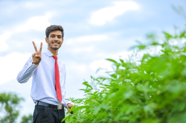 Joven agrónomo indio de pie en el campo y haciendo el signo de la victoria