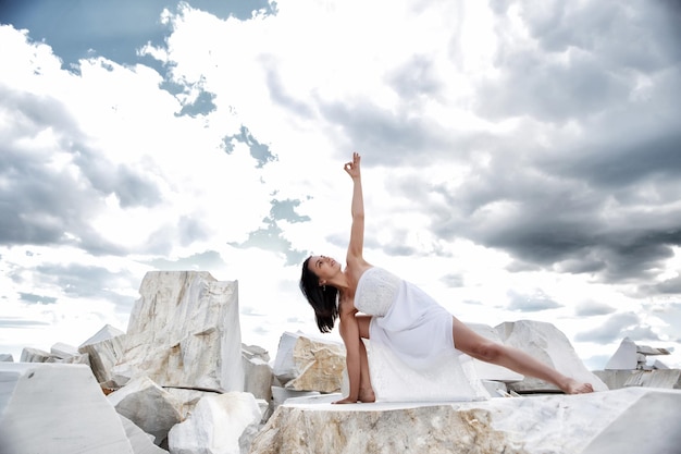 Una joven al aire libre parada en una pose de yoga con un largo vestido blanco contra el cielo azul