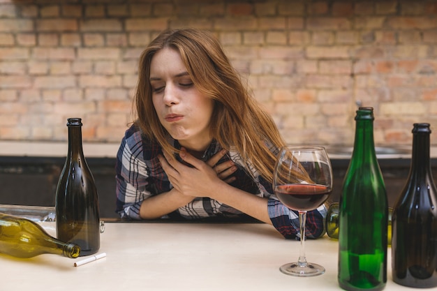 Joven alcohólica triste y perdida sentada en el sofá de la cocina bebiendo vino tinto, completamente borracha, deprimida, solitaria y sufriendo resaca en el alcoholismo y el abuso del alcohol.