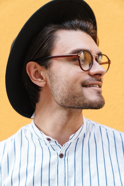 Foto joven alegre con anteojos y sombrero negro sonriendo y mirando a un lado en copyspace aislado sobre pared amarilla