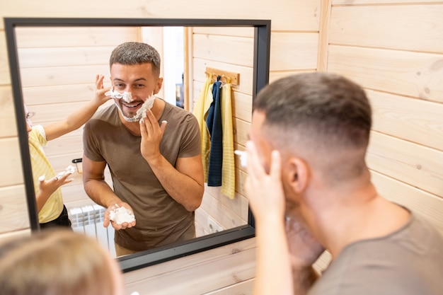 Joven alegre aplicando espuma en la barba mientras su linda hijita lo ayuda durante la higiene matutina, ambos de pie frente al espejo