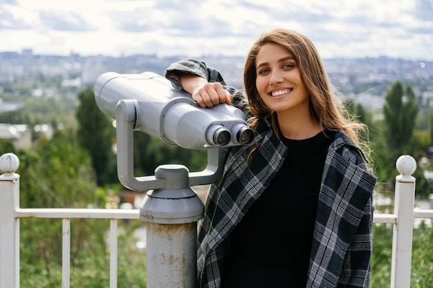 Una joven alegre se apoya en grandes binoculares mientras sonríe a la cámara