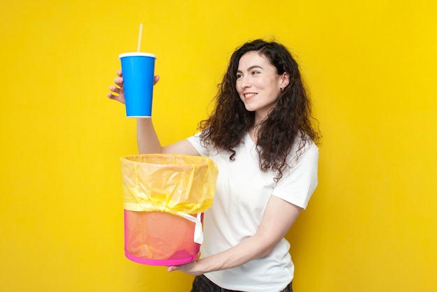 Una joven alegre arroja un vaso de plástico a un bote de basura, una mujer tira basura