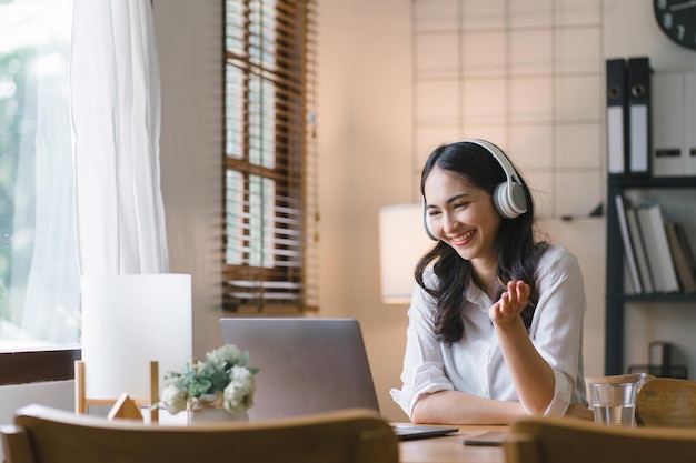 Una joven alegre con auriculares está teniendo una conferencia web en su mesa de la cocina mientras trabaja remotamente desde casa