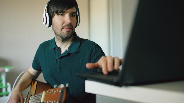 Foto un joven alegre con auriculares sentado en la cocina aprendiendo a tocar la guitarra usando una laptop