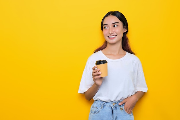 Una joven alegre y bonita con camiseta blanca sosteniendo una taza de café y mirando hacia otro lado en un espacio en blanco aislado en un fondo amarillo