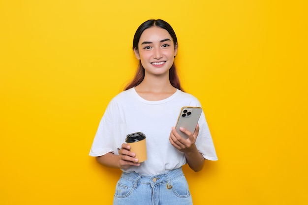 Una joven alegre y bonita con camiseta blanca sosteniendo un teléfono móvil y una taza de café aislada de fondo amarillo