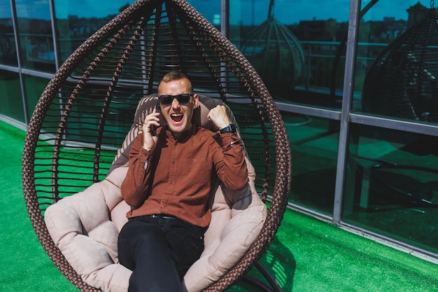 Un joven alegre con una camisa con un teléfono se sienta en la terraza de un edificio de oficinas después de que el trabajo usa internet inalámbrico. Un empleado positivo con anteojos está hablando por teléfono.