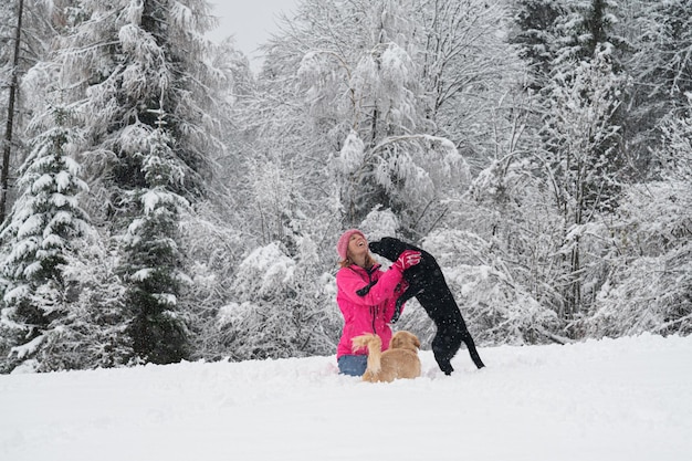 Una joven alegre con una chaqueta de invierno rosa sentada en la nieve jugando con sus dos perros.