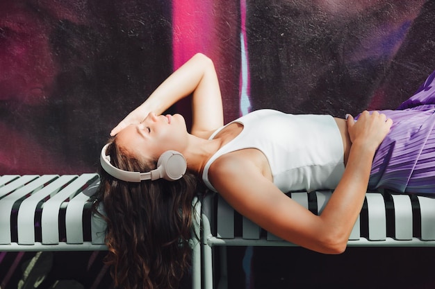 Una joven alegre está sentada en un banco con auriculares y escuchando música. Una hermosa niña disfruta de la música en la generación z de la ciudad.