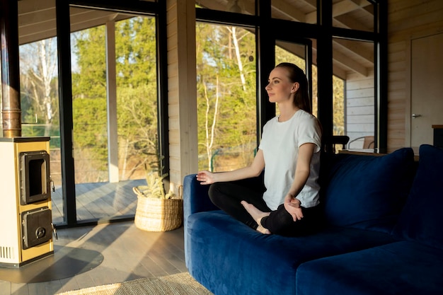 Una joven alegre está sentada en casa en un cómodo sofá azul y meditando una mujer hace yoga i