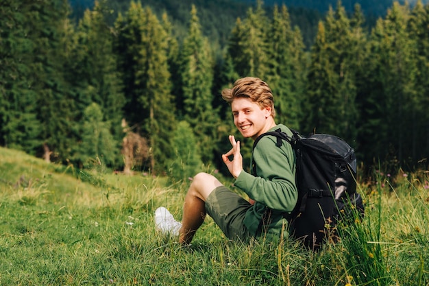 Un joven y alegre excursionista descansando en la hierba de las montañas y mostrando un gesto correcto con los dedos