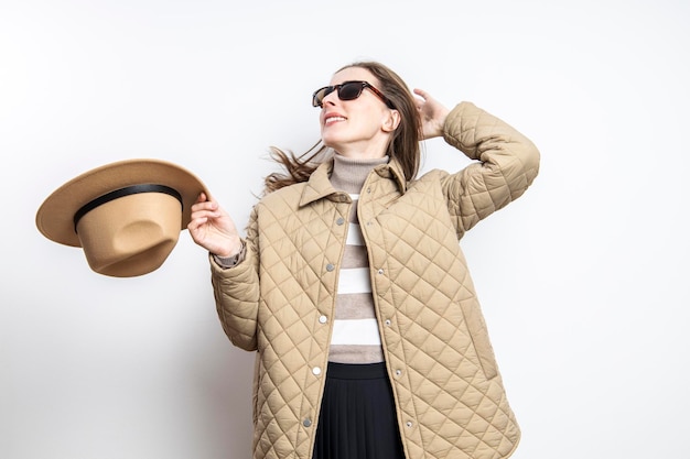 Una joven alegre con gafas y una chaqueta se quitó el sombrero cerca de una pared blanca