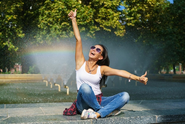 Una joven alegre con gafas de sol se sienta contra una fuente de fondo con arco iris