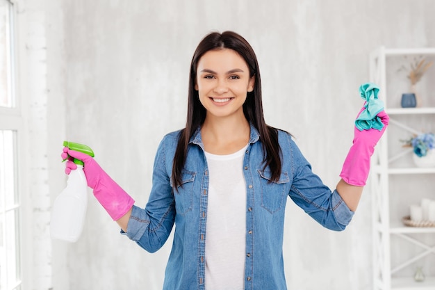 Una joven alegre con guantes de goma rosas usando un agente de limpieza y un limpiador de polvo
