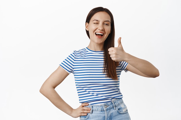 Joven alegre guiña un ojo y muestra los pulgares hacia arriba, sonriendo complacida, satisfecha con algo bueno, elogia el buen trabajo, cumplidos, de pie en una camiseta contra la pared blanca