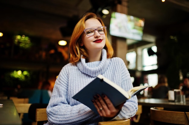 Una joven alegre y hermosa mujer pelirroja con gafas sentada en su lugar de trabajo en un café leyendo algo en su cuaderno