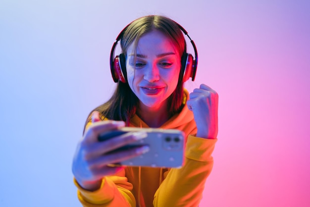 Una joven alegre juega juegos móviles con auriculares celebrando una victoria bajo un neón colorido