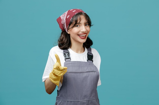 joven y alegre limpiadora que usa pañuelo uniforme y guantes de goma mirando y apuntando a la cámara aislada en el fondo azul