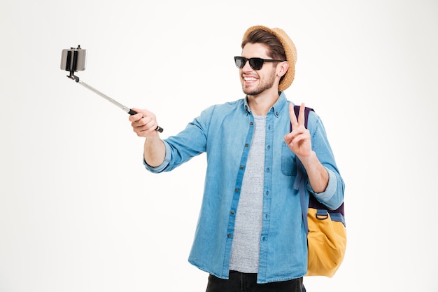 Joven alegre con mochila tomando fotografías con teléfono móvil y selfie stick