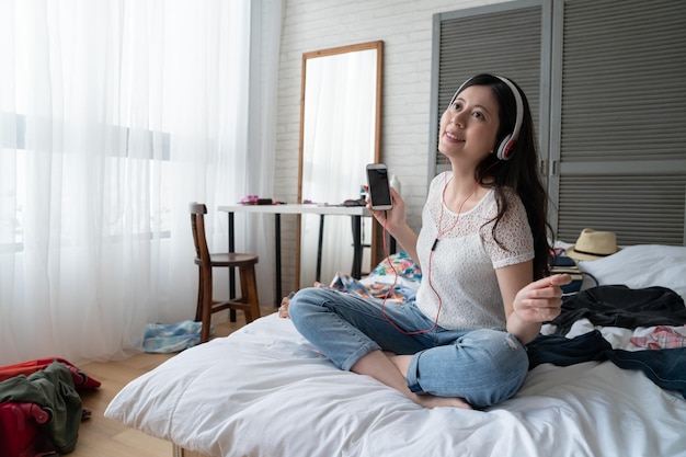 Joven alegre mujer asiática coreana sentada en la cama barajando canciones en el teléfono móvil. Chica relajada escuchando música con auriculares en el dormitorio de casa. Mujer feliz en casual disfruta del tiempo libre mientras empaca equipaje