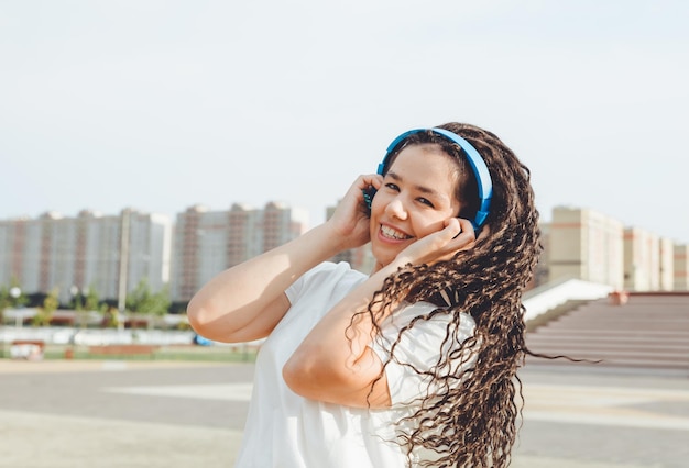 Una joven alegre mujer feliz con rastas vestida con una camiseta blanca bailando escuchando música con auriculares descansando relajándose en un parque de la ciudad caminando por un callejón Concepto de estilo de vida urbano
