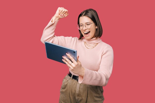 Joven alegre mujer de moda sosteniendo tableta y celebrando sobre fondo rosa