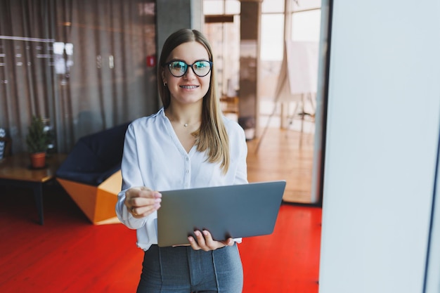 Una joven y alegre mujer de negocios de aspecto europeo con un pantalones blanco y anteojos sostiene una laptop portátil en sus manos cerca de una gran ventana en la oficina en el último piso