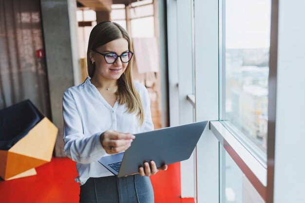 Una joven y alegre mujer de negocios de aspecto europeo con un pantalones blanco y anteojos sostiene una laptop portátil en sus manos cerca de una gran ventana en la oficina en el último piso